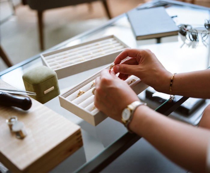 A person’s hands delicately pick up a gold ring from a velvet-lined jewellery tray on a glass table