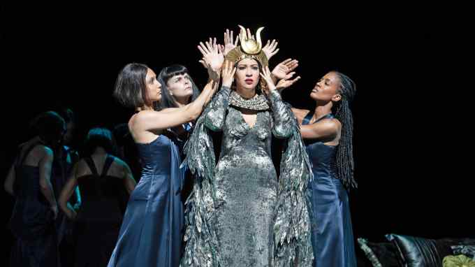 A woman in a silvery blue gown clutches her golden headpiece — other women in darker blue gowns stand around her with hands raised