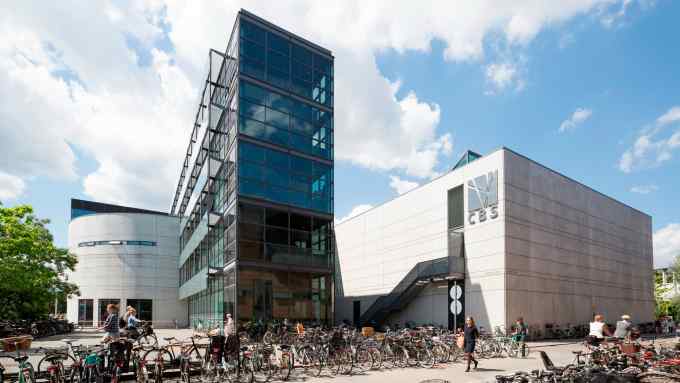 The exterior of Copenhagen Business School with its distinctive glass and concrete architecture. A busy bike parking area is in the foreground, with students and faculty walking by