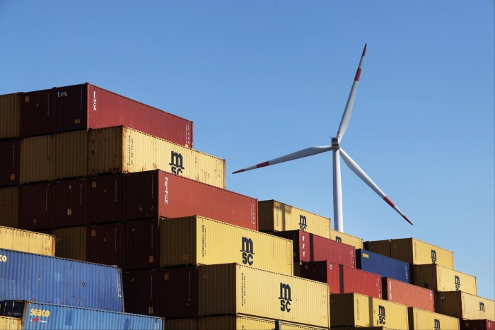 A wind turbine spins behind shipping containers stacked at Hamburg Port