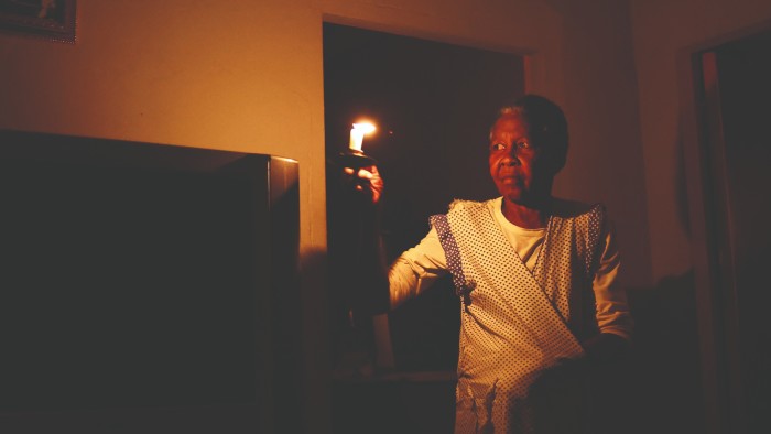 A person in a patterned dress holds a candle, illuminating a dark room with a soft, flickering light, highlighting a television and framed picture on the wall