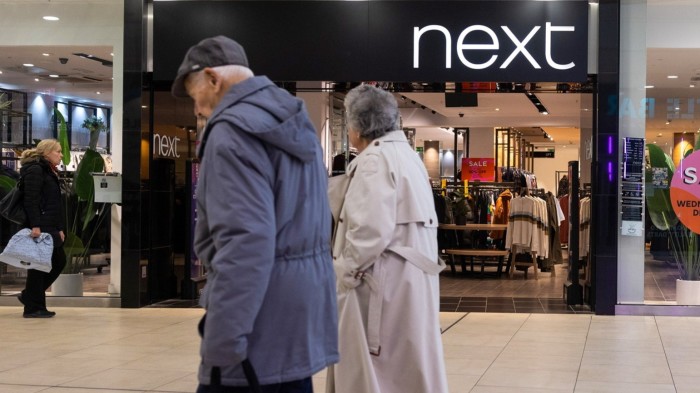 An exterior view of a Next clothing store in a shopping mall, with sale signs visible inside. Several people are walking by, including an elderly couple