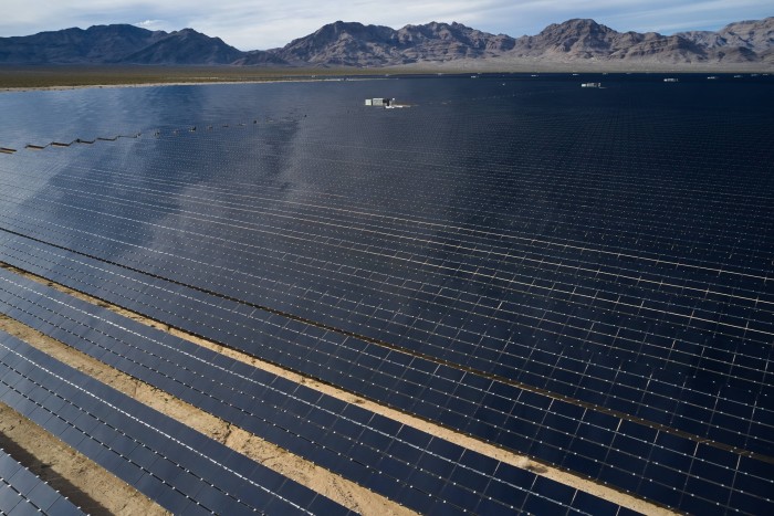 A large-scale solar farm with numerous dark solar panels arranged in rows, stretching across a desert landscape with mountains in the background