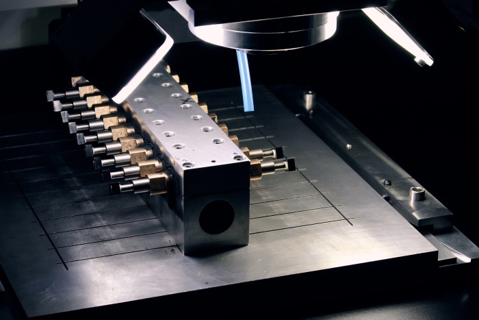 Placed at the centre of a laboratory environment is a metallic component featuring several small cylindrical extensions arranged in a structured and symmetrical pattern
