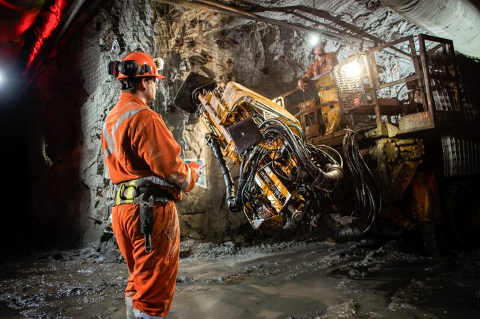 In a mining operation underground, a worker appears to observe or communicate with another worker who is operating a large mining equipment