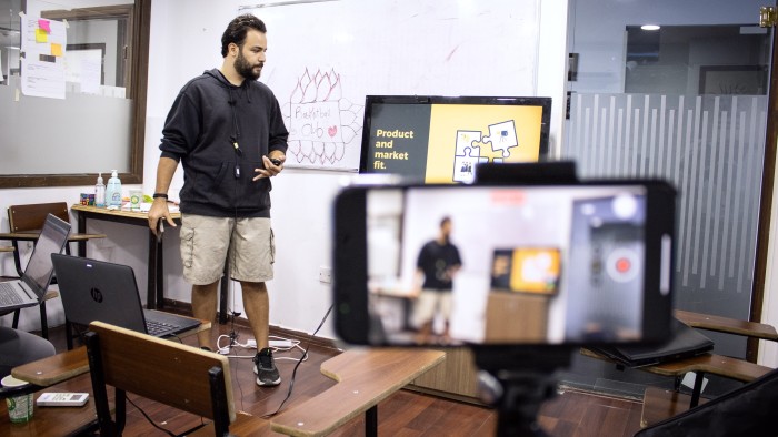 A man stands in front of a whiteboard giving a presentation, recorded by a smartphone on a tripod. The whiteboard includes a drawing and text about “Product and market fit.”