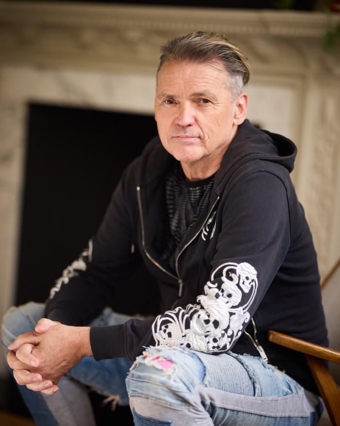 A man with short hair with a slightly swept-back style, showing streaks of natural greying. He’s wearing a zip-up hoodie with a printed shirt underneath